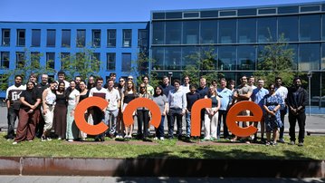 Gruppensfoto von Mitarbeitern vor einem großen, orangefarbenen Schriftzug "core" in einem modernen, hellen Gebäude.