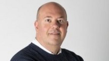 Ein Mann mit kurzen Haaren trägt einen dunkelblauen Pullover und steht mit verschränkten Armen vor einem neutralen Hintergrund. Er blickt direkt in die Kamera.