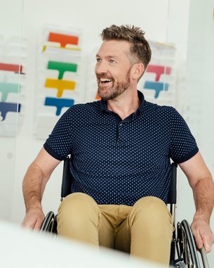 Ein Mann mit Bart sitzt in einem Rollstuhl und lächelt. Im Hintergrund sind bunte Farbmuster sichtbar.