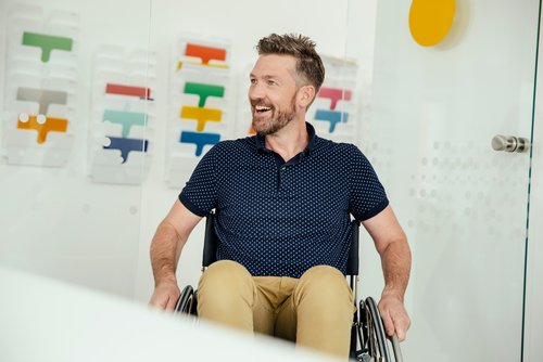 Ein Mann mit Bart sitzt in einem Rollstuhl und lächelt. Im Hintergrund sind bunte Farbmuster sichtbar.