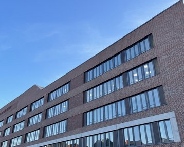 Moderne Backsteinfassade eines mehrstöckigen Gebäudes mit großen Fenstern und klaren Linien, unter einem blauen Himmel.