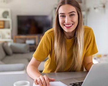 Eine lächelnde Frau mit langen Haaren sitzt an einem Tisch und arbeitet an einem Laptop. Sie trägt ein gelbes T-Shirt und hat eine Tasse neben sich. Im Hintergrund ist ein Wohnzimmer mit einem Sofa und einem Fernseher sichtbar.