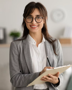 Eine Frau mit Brille trägt einen grauen Blazer und hält ein Notizbuch in der Hand. Sie steht in einem modernen Raum mit neutralen Farben und lächelt freundlich. Im Hintergrund sind Möbel und Pflanzen sichtbar.