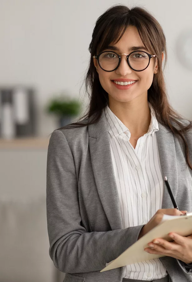 Eine Frau mit Brille trägt einen grauen Blazer und hält ein Notizbuch in der Hand. Sie steht in einem modernen Raum mit neutralen Farben und lächelt freundlich. Im Hintergrund sind Möbel und Pflanzen sichtbar.