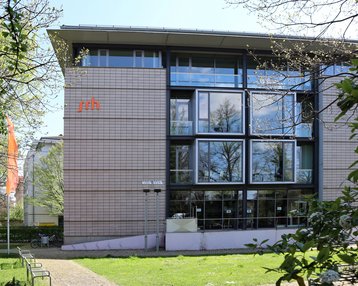 Moderne Gebäude mit großen Fenstern, beschriftet mit "srh", umgeben von Bäumen und Grünflächen.
