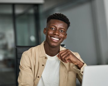 Ein junger Mann sitzt an einem Schreibtisch, lächelt und arbeitet an einem Laptop. Auf dem Tisch liegt ein Smartphone und einige Dokumente.