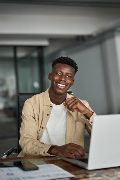 Ein junger Mann sitzt an einem Schreibtisch, lächelt und arbeitet an einem Laptop. Auf dem Tisch liegt ein Smartphone und einige Dokumente.
