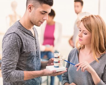 Ein Mann und eine Frau diskutieren über ein Modell der Wirbelsäule in einem Klassenzimmer. Im Hintergrund sind weitere Personen und anatomische Modelle sichtbar.