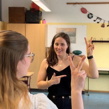 Eine Frau zeigt mit den Händen Zeichen und lächelt, während sie mit einer anderen Person in einem Klassenzimmer kommuniziert.