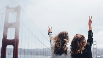 Two people are standing with their backs to the camera, making a peace sign with both hands. In the background, the Golden Gate Bridge and a cloudy sky can be seen.
