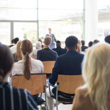 Eine Gruppe von Personen sitzt in einem Konferenzraum und hört einem Redner zu, der vorne spricht.