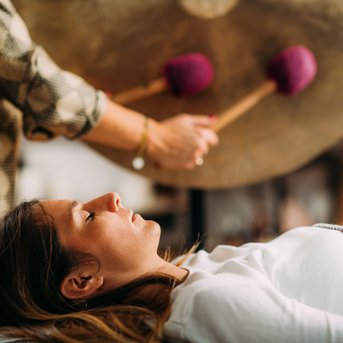 Eine Person liegt entspannt auf einer Liege, während eine andere Person mit zwei Schlägeln auf einem Gong spielt. Der Fokus liegt auf der Person, die auf der Liege liegt.