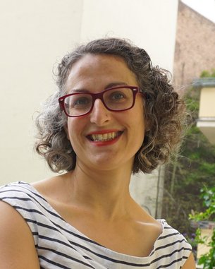 Eine Frau mit lockigem Haar und roten Brillenläsern lächelt freundlich. Sie trägt ein gestreiftes Oberteil und steht vor einer hellen Wand.