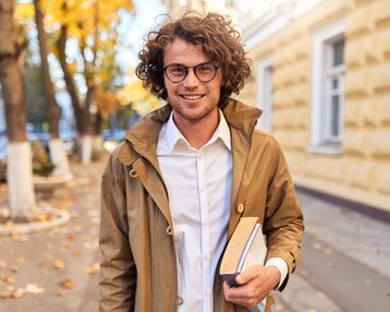Ein junger Mann mit lockigem Haar und Brille trägt eine beige Jacke und hält zwei Bücher in der Hand. Er steht auf einem Bürgersteig, umgeben von herbstlichen Bäumen und einem Gebäude im Hintergrund.