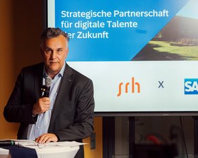Ein Mann steht an einem Tisch und hält ein Mikrofon, während im Hintergrund eine Präsentation zur strategischen Partnerschaft zwischen SRH und SAP zu sehen ist.