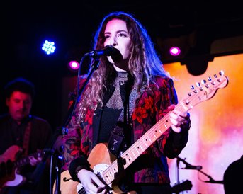 A musician with an electric guitar sings on stage while another musician plays in the background. Colorful lights illuminate the scene, creating a vibrant atmosphere.