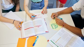 Several people are working at a table, surrounded by books, notes, and colorful markers, as they collaborate on a project or presentation.