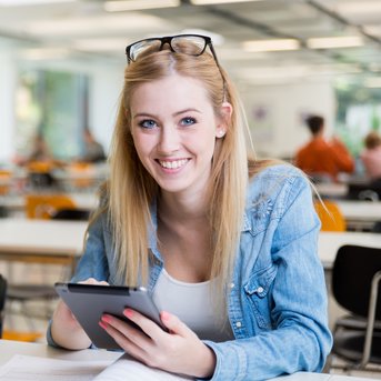 Eine junge Frau mit blonden Haaren sitzt an einem Tisch, hält ein Tablet und lächelt. Neben ihr steht eine Kaffeetasse.