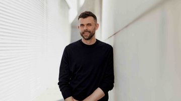 Ein Mann mit Bart steht lächelnd an einer Wand, gekleidet in einem schwarzen Pullover und beigen Hosen. Die Umgebung ist modern und minimalistisch, mit hellen Farben und Linien.