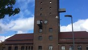 Eine Gruppe von Menschen steht vor einem hohen, rechteckigen Gebäude mit einer markanten Uhr und einem modernen Anbau. Der Himmel ist blau mit einigen Wolken, und Bäume sind im Vordergrund sichtbar.