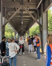 Eine Gruppe von Menschen steht unter einer Brücke, während eine Frau mit einem Fahrrad im Mittelpunkt steht. Einige Personen tragen Sicherheitswesten, und es gibt eine Diskussion oder Präsentation.