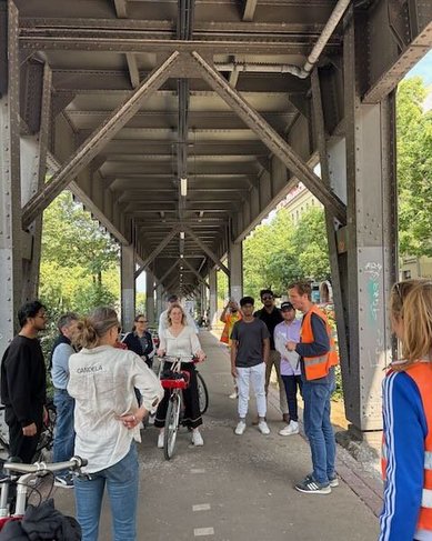 Eine Gruppe von Menschen steht unter einer Brücke, während eine Frau mit einem Fahrrad im Mittelpunkt steht. Einige Personen tragen Sicherheitswesten, und es gibt eine Diskussion oder Präsentation.