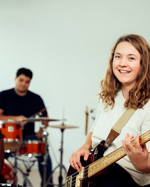 Eine Frau mit einer Bassgitarre lächelt in die Kamera, während sie in einer Bandprobe mit anderen Musikern spielt.