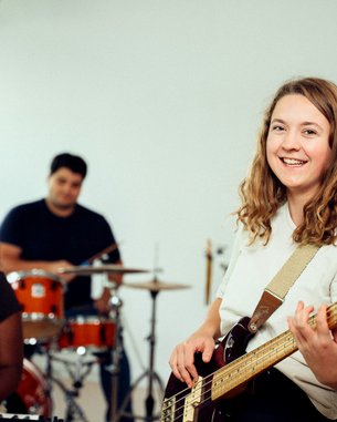 A woman with a bass guitar smiles at the camera while playing in a band rehearsal with other musicians.
