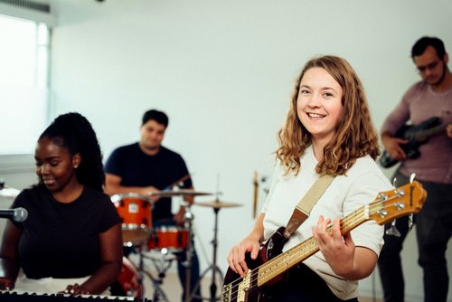 Eine Frau mit einer Bassgitarre lächelt in die Kamera, während sie in einer Bandprobe mit anderen Musikern spielt.