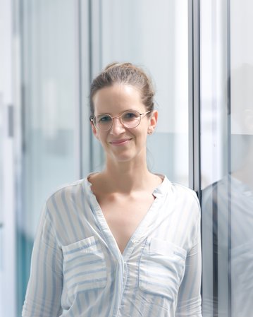 Eine Frau mit Brille steht in einem modernen Flur, lächelt und trägt ein gestreiftes Hemd. Helle, minimalistische Umgebung.