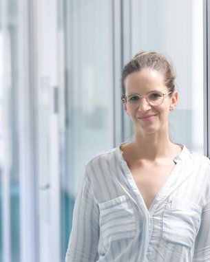 A woman with glasses stands in a modern hallway, smiling and wearing a striped shirt. The environment is bright and minimalist.