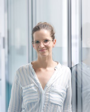 Eine Frau mit Brille steht in einem modernen Flur, lächelt und trägt ein gestreiftes Hemd. Helle, minimalistische Umgebung.