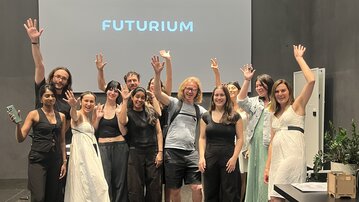 A group of twelve people stands in a room, raising their hands. In the background, there is a screen with the inscription 'FUTURIUM'.