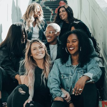 Six women are sitting on a colorful staircase, smiling and chatting. They are dressed casually and have different hairstyles.