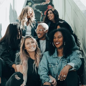 Sechs Frauen sitzen auf einer bunten Treppe, lächeln und unterhalten sich. Sie tragen lässige Kleidung und haben unterschiedliche Frisuren.