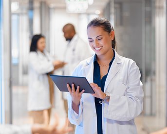 Eine Ärztin in einem weißen Kittel hält ein Tablet und lächelt, während sie in einem modernen Krankenhausflur steht. Im Hintergrund sind weitere medizinische Fachkräfte zu sehen.