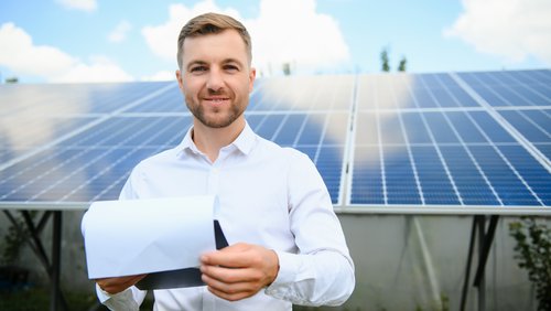 Mann in weißem Hemd hält ein Dokument vor einer Solaranlage. Im Hintergrund sind mehrere Solarpanels sichtbar.