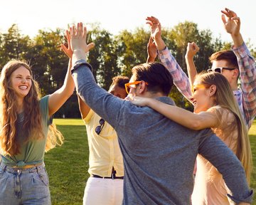 A group of young people is celebrating outdoors, raising their hands and smiling. They are standing in a meadow, surrounded by trees.