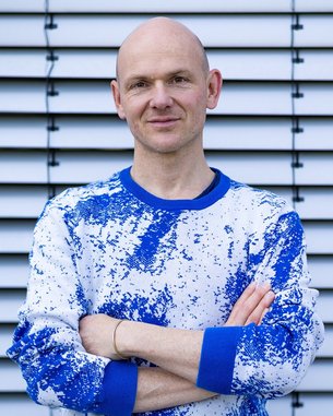 Ein Mann mit Glatze trägt einen blauen und weißen Pullover und steht mit verschränkten Armen vor einer gestreiften Wand.