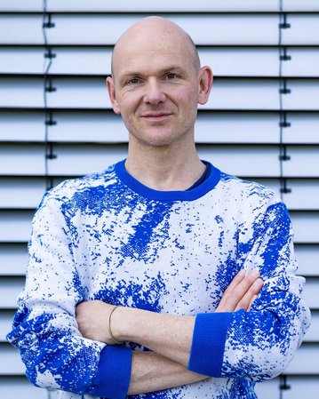 Ein Mann mit Glatze trägt einen blauen und weißen Pullover und steht mit verschränkten Armen vor einer gestreiften Wand.