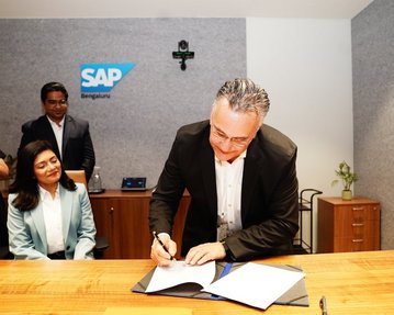 Ein Mann in einem Anzug unterschreibt ein Dokument an einem Tisch, während eine Frau und ein weiterer Mann im Hintergrund zuschauen. SAP-Logo sichtbar.