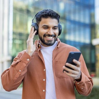 Man with headphones, smiling as he looks at his smartphone while walking past a modern building.
