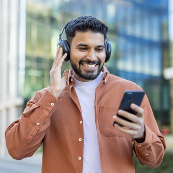 Mann mit Kopfhörern, der lächelnd auf sein Smartphone schaut, während er an einem modernen Gebäude vorbeigeht.