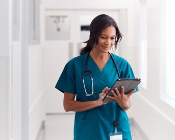 Eine Krankenschwester in grüner Uniform steht in einem Flur und betrachtet ein Tablet. Ein Stethoskop hängt um ihren Hals.