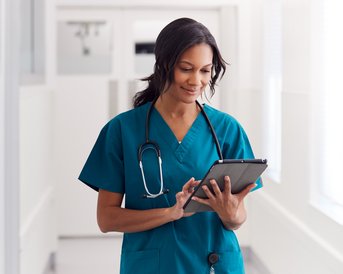 Eine Krankenschwester in grüner Uniform steht in einem Flur und betrachtet ein Tablet. Ein Stethoskop hängt um ihren Hals.