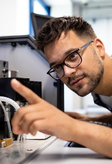 Ein Mann mit Brille untersucht eine Maschine, während er mit dem Finger auf ein Detail zeigt.