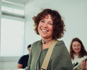 Eine Frau mit lockigem Haar spielt Bass in einer Bandprobe, lächelnd und in einem lässigen Outfit. Im Hintergrund sind Schlagzeug und andere Musiker sichtbar.