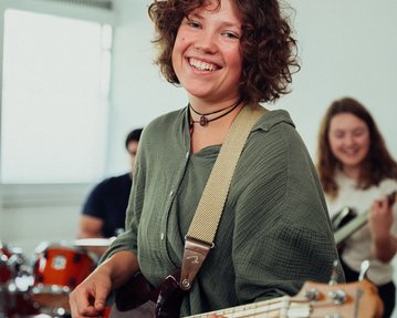 Eine Frau mit lockigem Haar spielt Bass in einer Bandprobe, lächelnd und in einem lässigen Outfit. Im Hintergrund sind Schlagzeug und andere Musiker sichtbar.