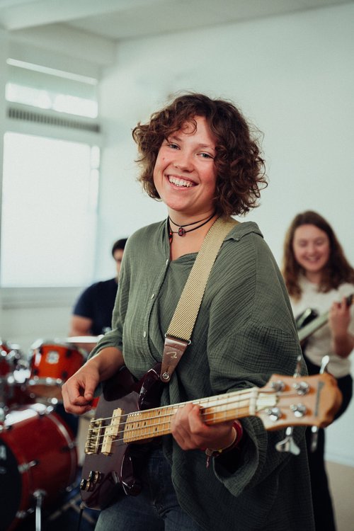 Eine Frau mit lockigem Haar spielt Bass in einer Bandprobe, lächelnd und in einem lässigen Outfit. Im Hintergrund sind Schlagzeug und andere Musiker sichtbar.