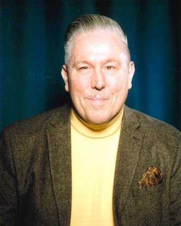 Ein Mann mit grauen Haaren trägt einen braunen Blazer und ein gelbes Oberteil. Er lächelt in die Kamera vor einem blauen Hintergrund.